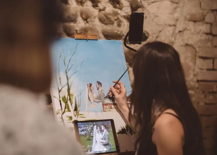 An artist paints a wedding ceremony scene at an easel, with a reference photograph below.